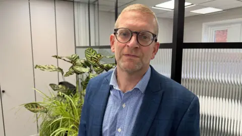 MoJ owes us £20m after contractor ISG's collapse, say suppliers Ian standing in his office. He is wearing a smart blue suit with a blue and white shirt. He is standing in front of a big green plant. There is a board room behind him but we can't see it because the glass is frosted.
