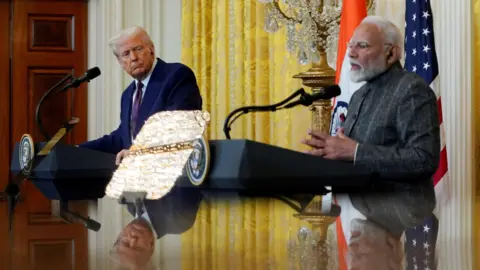 Trump says Modi has agreed to stop buying Russian oil Reuters Two men sit in front of a reflective table with American and Indian flags in the background.