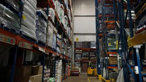 NI customers paying more post-Brexit, says catering firm boss A warehouse with shelves of stock. There is a concrete floor with yellow markings.