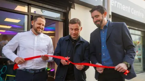 Children from Belfast low income families to be given free tuition PA Media Three men are holding a red ribbon outside a building. The man in the middle is cutting the ribbon with scissors.