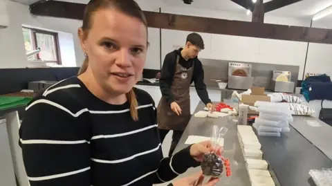 Kent chocolatier selling smaller bars amid cocoa price surge BBC/LIZ SAUL A woman in a stripey navy and white jumper, with long light brown hair, tied in a pony tail. She is standing in the kitchen of the chocolate factory, holding a clear bag of chocolate nuts. In the back ground, a young man is folding gift boxes from cardboard templates.