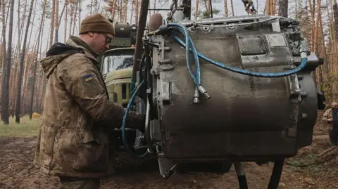 What do US sanctions against Russian oil companies mean for the war in Ukraine? Getty Images A man in military garb, beanie and sunglasses, with a Ukraine flag badge on his jacket sleeve, tends to a large piece of machinery in the middle of the woods