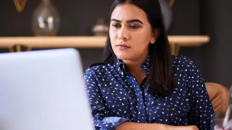 Getty Images A woman in a blue shirt sitting at a desk looking at a laptop.