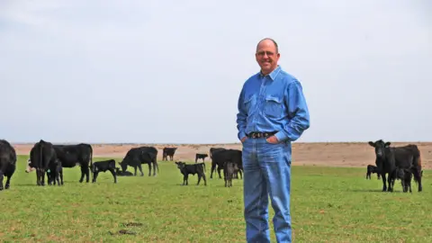 US beef prices are soaring. Will Trump's plans lower them? Mike Callicrate A man wearing denim stands on a grassy plot of land with his hands in his pockets. Several cows graze the land in the background.