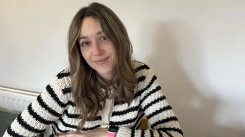 When should you turn the heating on? Abbi Lily Abbi, a young woman with dark blonde hair. She is wearing a black and white striped cardigan and is sitting at her kitchen table