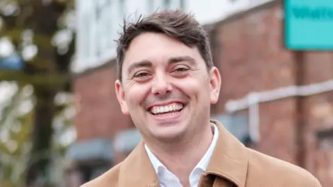 Essex inventor up for Earthshot Prize for tackling microplastics Matter Adam Root is wearing a beige jacket over a white shirt. He is standing in front of a brick building and smiling. He has short dark hair.