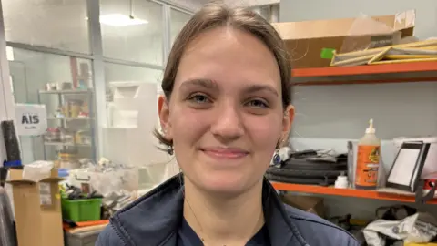BBC Millie Brown, a young lady with brown hair in a ponytail, smiles from inside an engineering lab at her university. There are busy-looking shelves and piles of equipment, boxes and other items behind her.