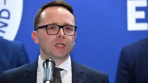 'Flawed' HMRC system stops hundreds of NI families' child benefit PA Media Dáire Hughes speaking at a microphone. He has dark hair and is wearing glasses. He has a navy suit and black tie.