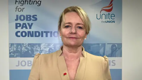 UK targets £420m at sky high industry energy bills Sharon Graham standing in front of a banner with Unite's branding on it - she is wearing a cream / gold structured top,  has blond hair, is wearing pearl-like earrings, has a poppy on her lapel, and has a neutral expression