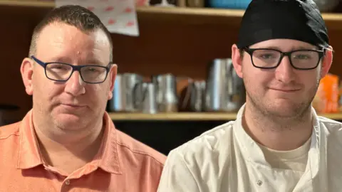 Workers back minimum wage rise 'as everything getting expensive' Ian and Jonathan Corlett standing next to each other, both wearing black rimmed glasses. Ian is wearing an orange T-shirt and Jonathan is wearing chefs whites and a black hat.