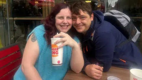 What went wrong with Pizza Hut? Joanne, left, smiling, wearing a sleeveless blue top with shoulder length wavy hair, holding a take-out beverage cup. Chris, right, smiles, and is wearing a dark blue top and a rucksack. He is leaned over with his face next to Joanne's.