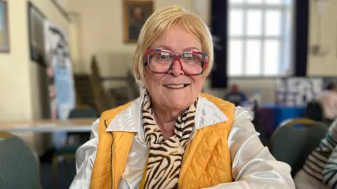 Norfolk council roadshow aims to help people with household bills Clare Worden/ BBC Hillary Letch has a blonde bob and red glasses. She is wearing a white shirt and yellow gilet.