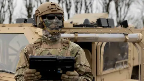 What's the best way to detect and destroy drones? MyDefence A soldier wearing a helmet, mask and sunglasses holds a large table computer and looks upwards. Behind him is a military vehicle.