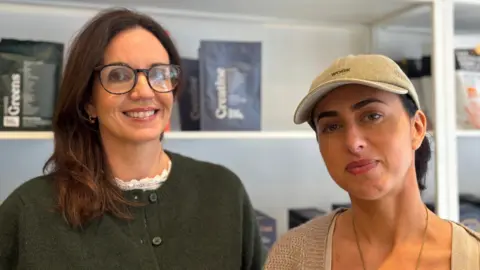 New businesses in Jersey's share shops to keep costs down Vanessa and Sophie are standing indoors in front of white shelving units displaying various packaged products. The shelves also feature framed artwork and decorative items. Vanessa is wearing a dark green buttoned cardigan with a white lace collar, and Sophie is wearing a light beige knit cardigan over a top, accessorized with a necklace and a light-colored cap.