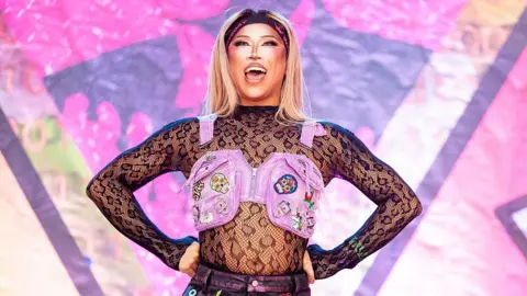 The fall and future of Manchester Pride Getty Images Drag queen Saki Yew is on-stage at Manchester Pride. She is wearing a lace top with long sleeves and a small pink waistcoat over the top of it, covered in brightly-coloured badges. She is open-mouthed, looking out from the stage. She is wearing a black headband with pink letters on it and has long, blonde hair.