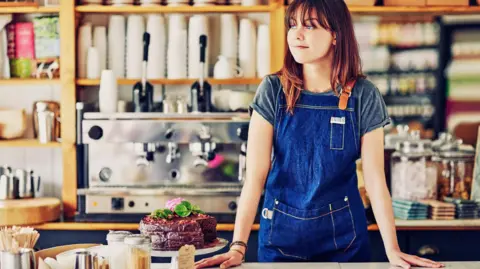 How much are the National Minimum Wage and National Living Wage worth? Getty Images A young woman with long reddish-brown hair and wearing a dark blue apron stands behind the counter in a brightly-lit cafe, next to an ornate chocolate cake. There is a large coffee machine on the back wall, with piles of empty take-away cups and several boxes of tea bags.
