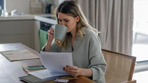 Why this month's inflation figure matters for you Getty Images A woman sits in a chair at home looking at paperwork and drinking a cup of tea