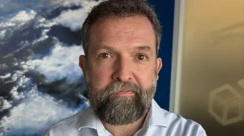 A man with grey hair and a grey beard looks into the camera wearing a neutral expression. He is wearing a white shirt and is standing in front of a photo of Earth taken from space.