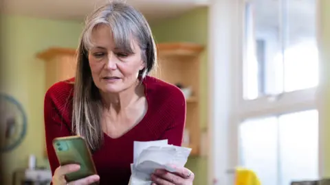 Why this month's inflation figure matters for you Getty Images Middle-aged woman with long grey hair looks at a pile of receipts and her mobile phone. She has a slightly anxious look on her face.