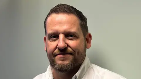 Children from Belfast low income families to be given free tuition Ciaran Toman is looking into the camera. He's wearing a white shirt and has brown hair. He's standing with a wall behind him.