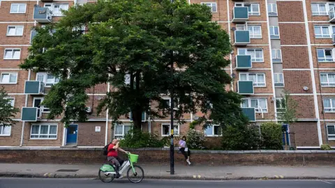 Families are priced out of London, Assembly told In Pictures via Getty Images A block of social housing flats viewed from the road, with a man cycling in front of them and a large leafy tree behind him, in front of the flats.