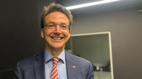 New businesses in Jersey's share shops to keep costs down Kirsten is dressed in formal attire, wearing a navy blue suit jacket, a light blue shirt, and a red-and-white striped tie. The background features a dark wall with a bright horizontal light fixture and a reflective surface, suggesting an indoor professional or studio environment