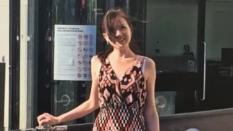 Contributed Marie Cattermole is a woman with brunette hair who is standing outside and leaning against a barrier. She is looking at the camera and smiling and is wearing a brown, orange and white patterned dress. 