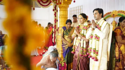 Janani Mohan/Yagappa Photography Janani Mohan and her husband in traditional Indian wedding clothing with garlands round their necks, stand with their hands in a prayer position in front of them, surrounded by guests celebrating their wedding