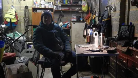 Essex inventor up for Earthshot Prize for tackling microplastics Adam Root Adam Root working in a dark garage surrounded by clutter. He is wearing a hat, coat and gloves and is sitting at a desk.