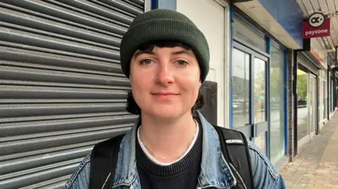 The new hubs saving Yorkshire towns from becoming banking deserts Kit Taylor/BBC A person smiling at the camera, wearing a dark green beanie hat, black jumper and a denim jacket. In the background are shut down shops.