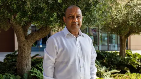 Getty Images Sanjay Shah standing outside underneath trees in what appears to be a garden in a built up office area - he is a middle aged man, with a white shirt and thinning hair. He is looking just past the camera with a neutral expression. 