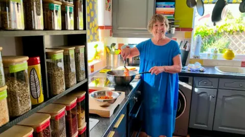 Positive Community Action celebrates 8,000 meals in Wiltshire Positive Community Action A woman in a blue dress cooks a meal in a pan in a tidy kitchen and smiles at the camera. In the foreground are several herbs and spices.