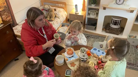 'I left Wales and moved to England for free childcare' A nursery worker wearing a red hoody is sitting around a low round table with four toddlers pretending to have a tea party. There is a small bed in the background with two teddy bears on it.