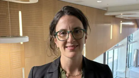 Lecturer Dr Christie Maddock smiles in a modern-looking building at Strathclyde Uni. She has glasses and brown hair, tied back. She wears a dark blazer, a green blouse and a green and gold beaded necklace.