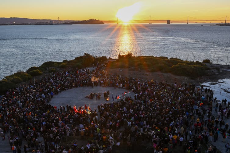 An Indigenous approach shows how changing the clocks for daylight saving time runs counter to human nature – and nature itself A group of people stand around an open circle on an island, as the Sun rises behind a bridge across the water.