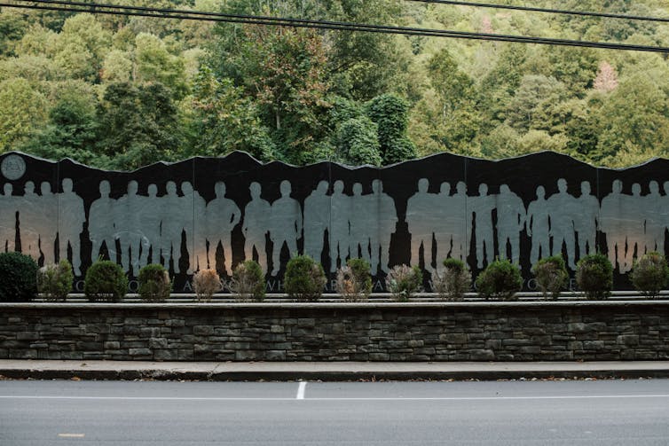 For many miners, religion and labor rights have long been connected in coal country A black wall with paler silhouettes of men standing along it, positioned above a low stone wall next to the road.