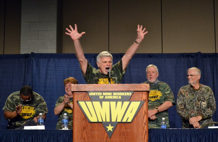 For many miners, religion and labor rights have long been connected in coal country A man with white hair raises his arms as he speaks animatedly at a podium, while he and other men nearby wear camo-colored t-shirts.