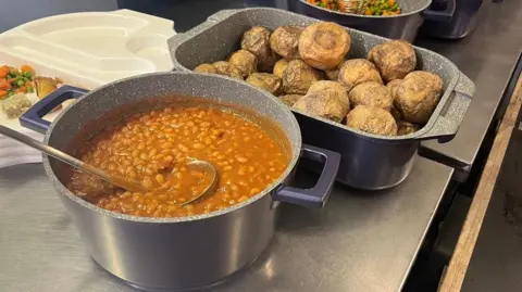 Devon to see 1,300 more children get free school meals A large pot of baked beans with a serving ladel and a large oven tin full of baked potatoes.