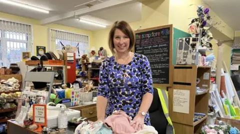 BBC A woman in a blue and black patterned dress in a room with boxes and shelves packed with cloth and other objects