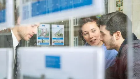 Getty Images A picture looking through an estate agent's window showing a young couple talking to an estate agent who is wearing a grey suit