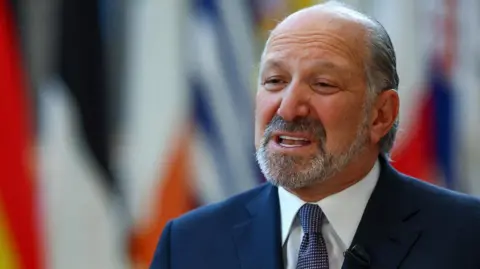 Reuters US Commerce Secretary Howard Lutnick wearing a navy blue suit, white shirt and blue dotted tie standing in front of a selection of out-of-focus country flags