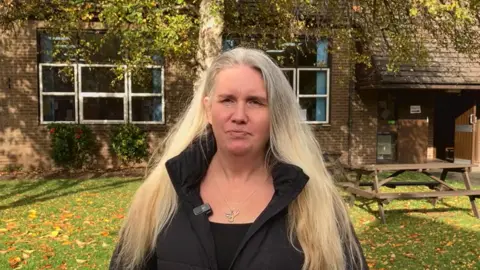 BBC A woman with long blonde hair is pictured outside a red brick building while wearing all black clothing. 
