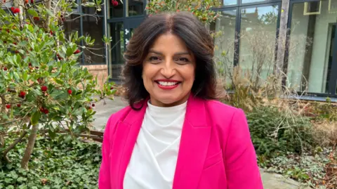 BBC Charnjit Saranna smiles at the camera. She is in front of a green tree with red berries and is wearing a pink jacket and white shirt.