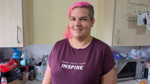 A woman with pink hair and wearing a burgundy t-shirt with 'dream, create, share, inspire' written across the front stands in a kitchen. Behind her are cabinets on the wall and a work surface. On the work surface is a microwave, a large bag of pasta, salt and pepper, measuring cups, a pot of utensils.