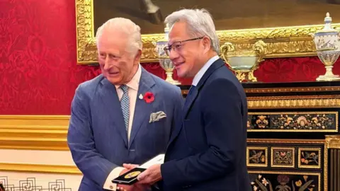 BBC King Charles handing a prize to Jensen Huang - the King is wearing a blue pinstripe suit and has a poppy on his lapel, Jensen Huang is holding the gold medal in its case and is wearing a dark suit and smiling with glasses on - the room they are in is very grand, with gold lining around a big mirror behind them, and red wall paper  