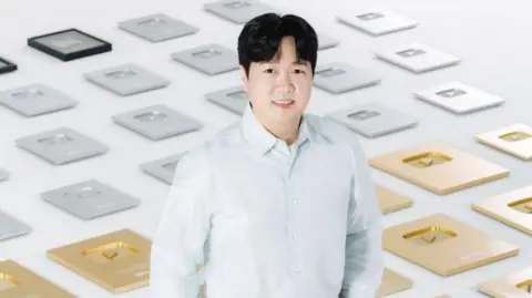 Courtesy of Pinkfong Pinkfong CEO Kim Min-seok standing at the centre of the frame with his hands in his pocket. Behind him is a series of gold- and silver-plated YouTube plaques.