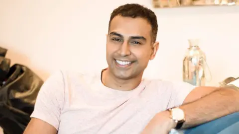 Jas Bains A man who appears to be in his 30s - he is sitting on a sofa at home, wearing a light off-white t-shirt, and smiling, clearly relaxed in the setting - he is leaning on the arm of the sofa with his left arm, and his watch is visible 