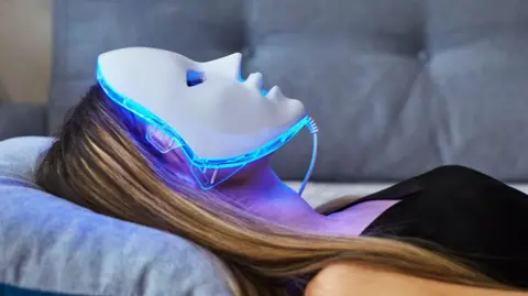 Getty Images A woman with long straight blonde hair lying flat on a blue sofa with an led mask on with blue light beaming on to her face