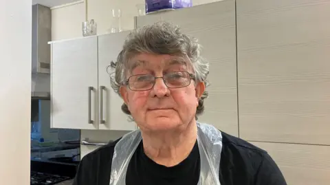 One in four missing meals due to cost pressures An older man with short grey hair and glasses. He is wearing a black t-shirt and a plastic apron and standing in a kitchen.