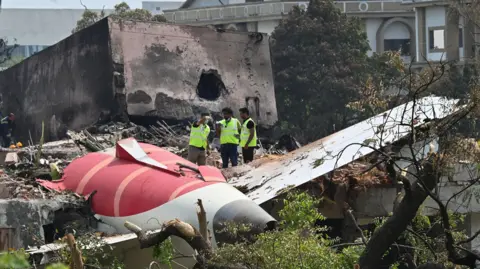 Getty Images The aftermath of the Air India crash showing a part of the plane crashed in the ground. Three men in high-vis jackets inspect the sight. Taken on 15th June 2025.
